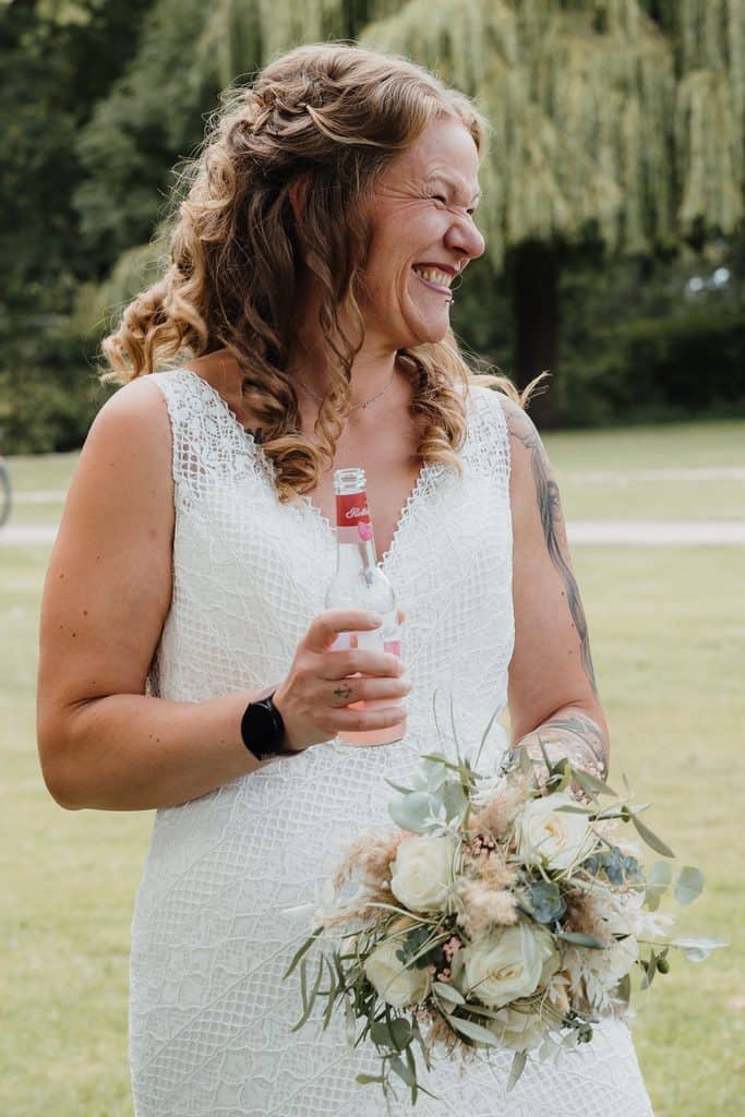 Lachende Braut mit Sektflasche und Brautstrauß bei einer unkonventionellen Hochzeit.
