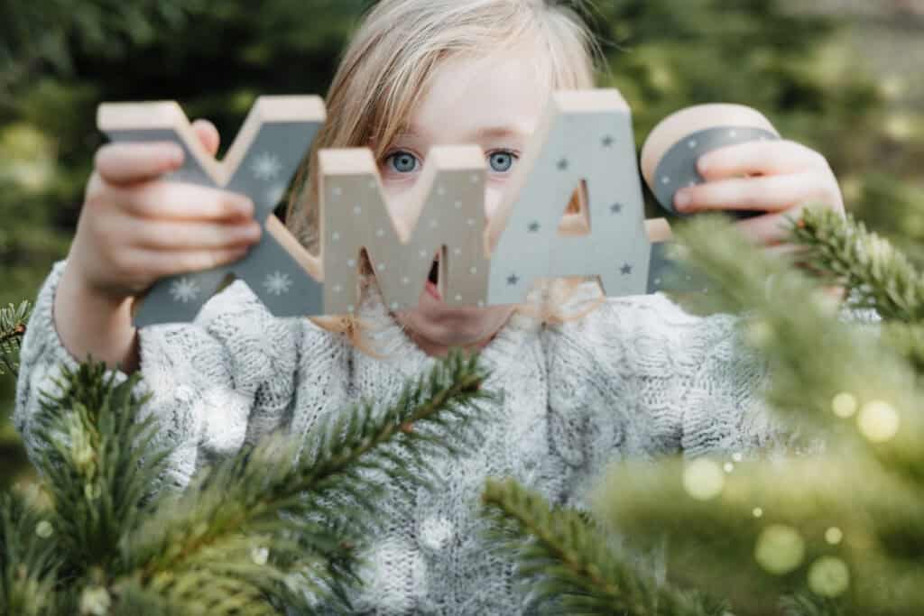 Kind zwischen Tannen hält weihnachtliche Holzbuchstaben in die Kamera